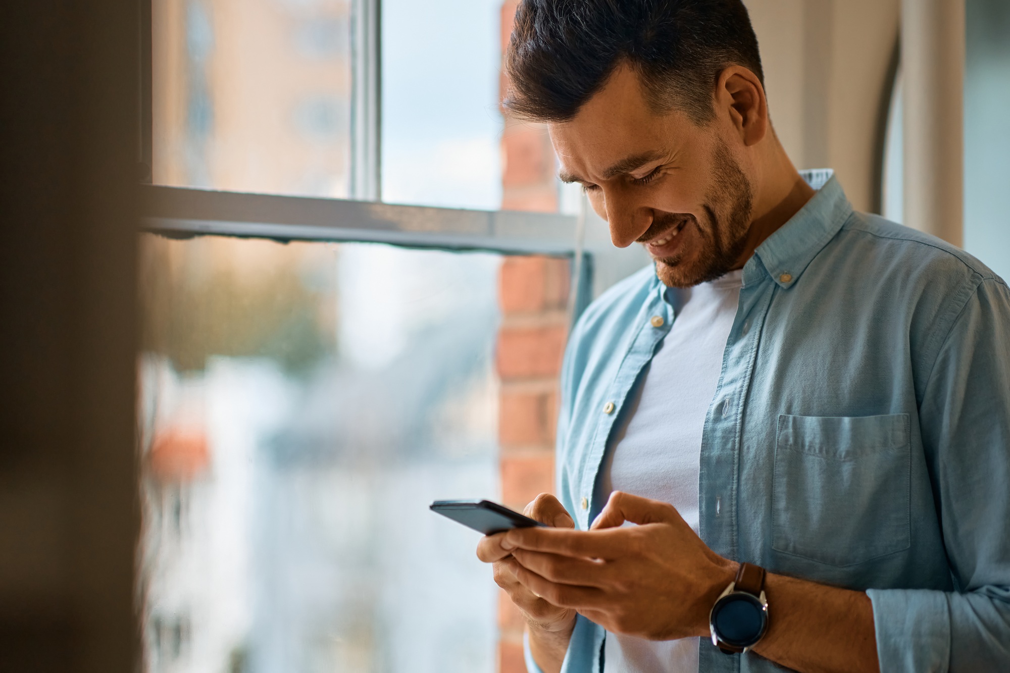 Happy man text messaging on smart phone by the window.
