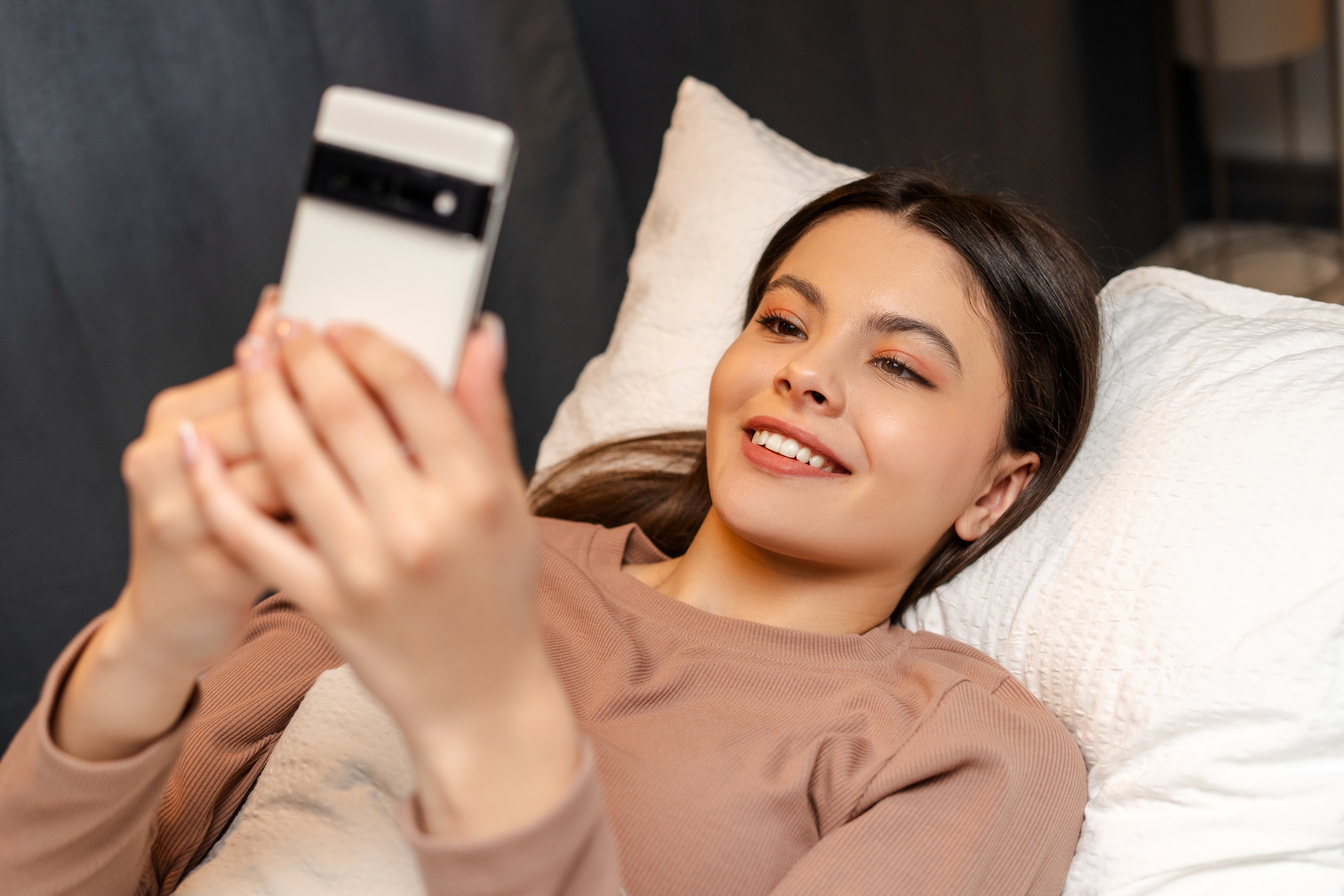 Happy young girl, teenager lying in bed using smartphone at night, text message