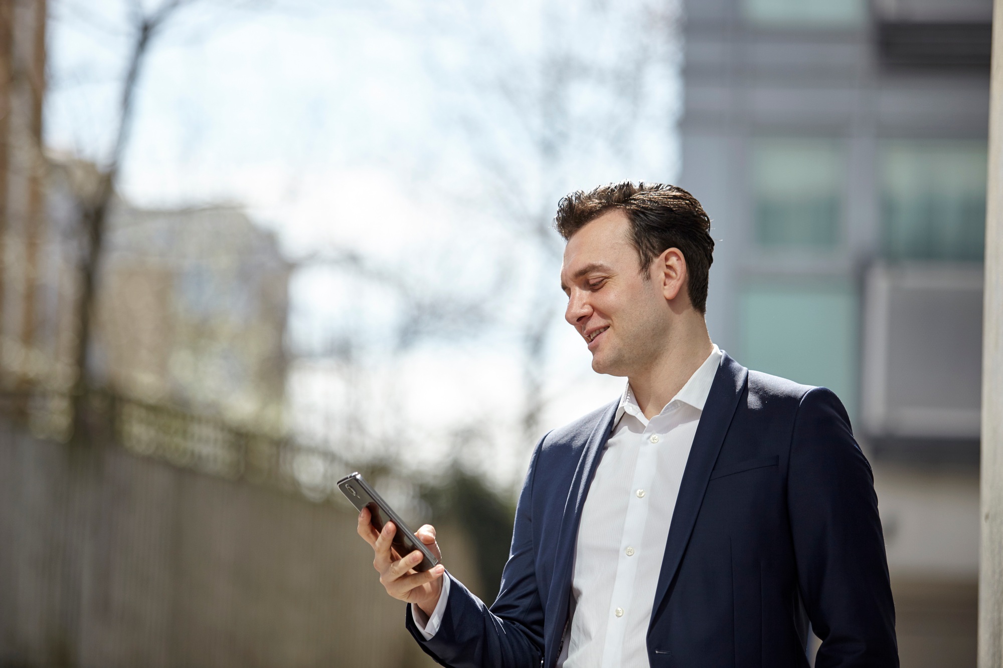 Businessman reading smartphone texts outside office, London, UK