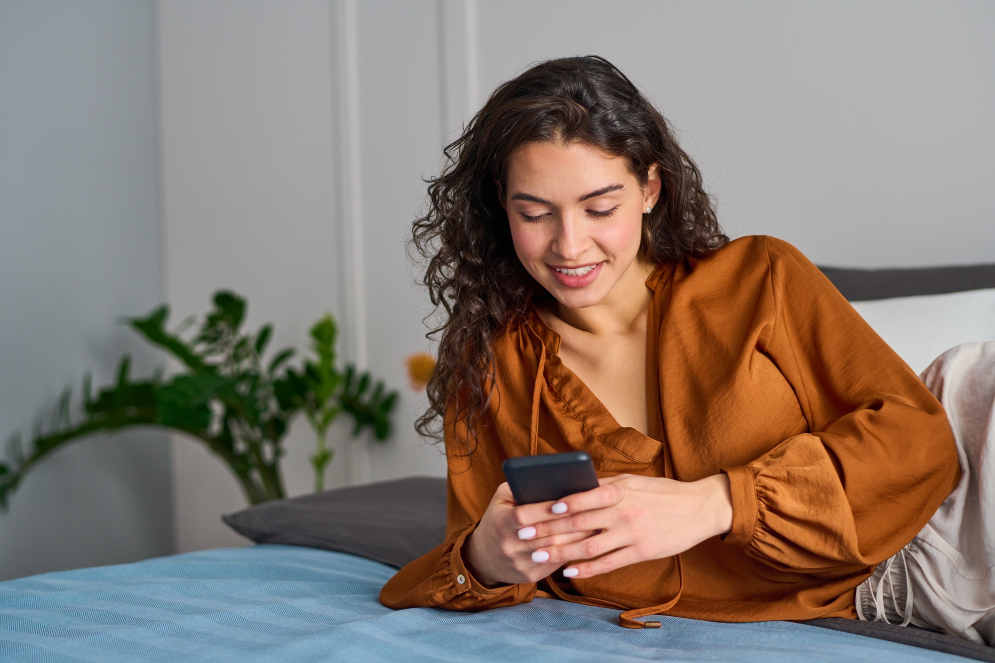 Smiling girl in silk blouse of bronze color lying on doubel bed and texting