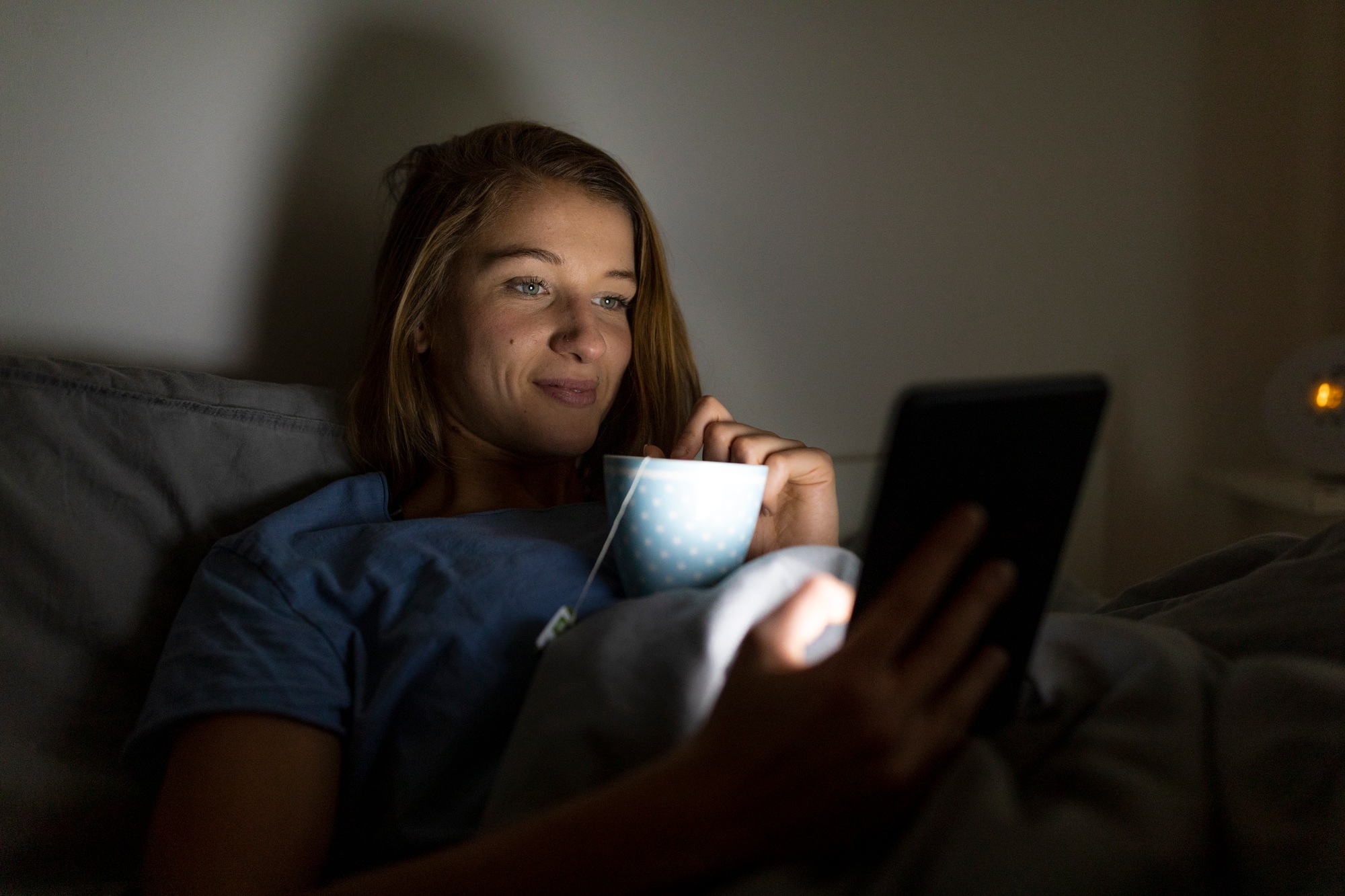 Young woman lying in bed at home with cup of tea at night using tablet